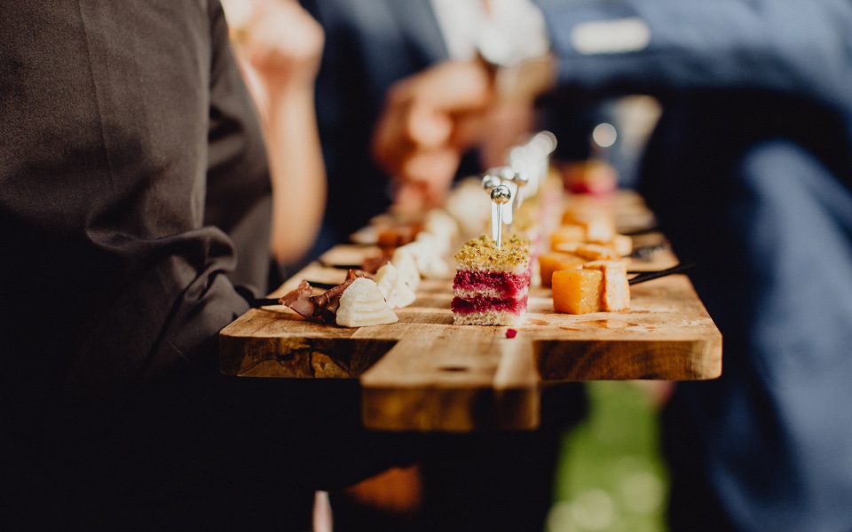 Plateau en bois de canapés gourmet colorés (bœuf, fromage, betterave) tenus par un serveur. Focus sur la petite pièce montée rose.