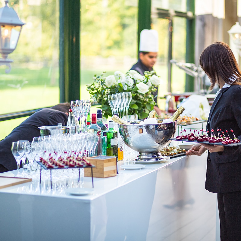 Bar de cocktail avec champagne et verrerie. Une serveuse en uniforme noir sert des canapés lors d'une réception traiteur élégante.
