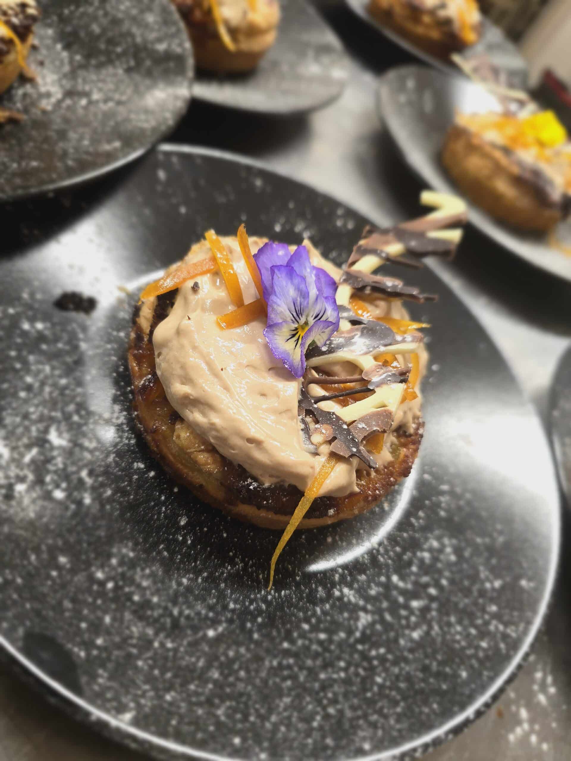 Pâtisserie ronde nappée de mousse au chocolat, zestes d'orange, chocolat et fleur comestible violette sur assiette noire.