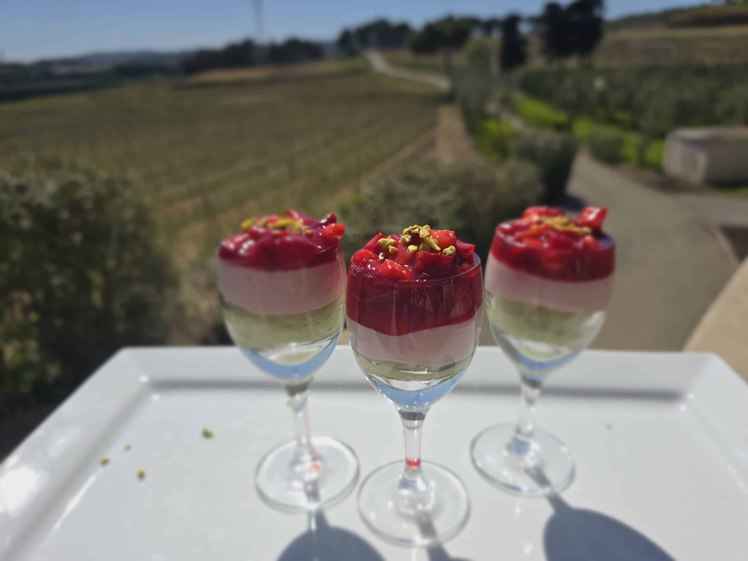 Verrines étagées aux fruits rouges et pistache sur un plateau blanc, offrant une vue imprenable sur un vignoble.